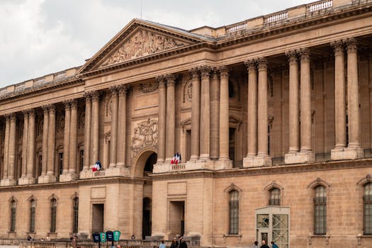 Front view of the iconic Louvre Museum facade, Paris, with French flags displayed.