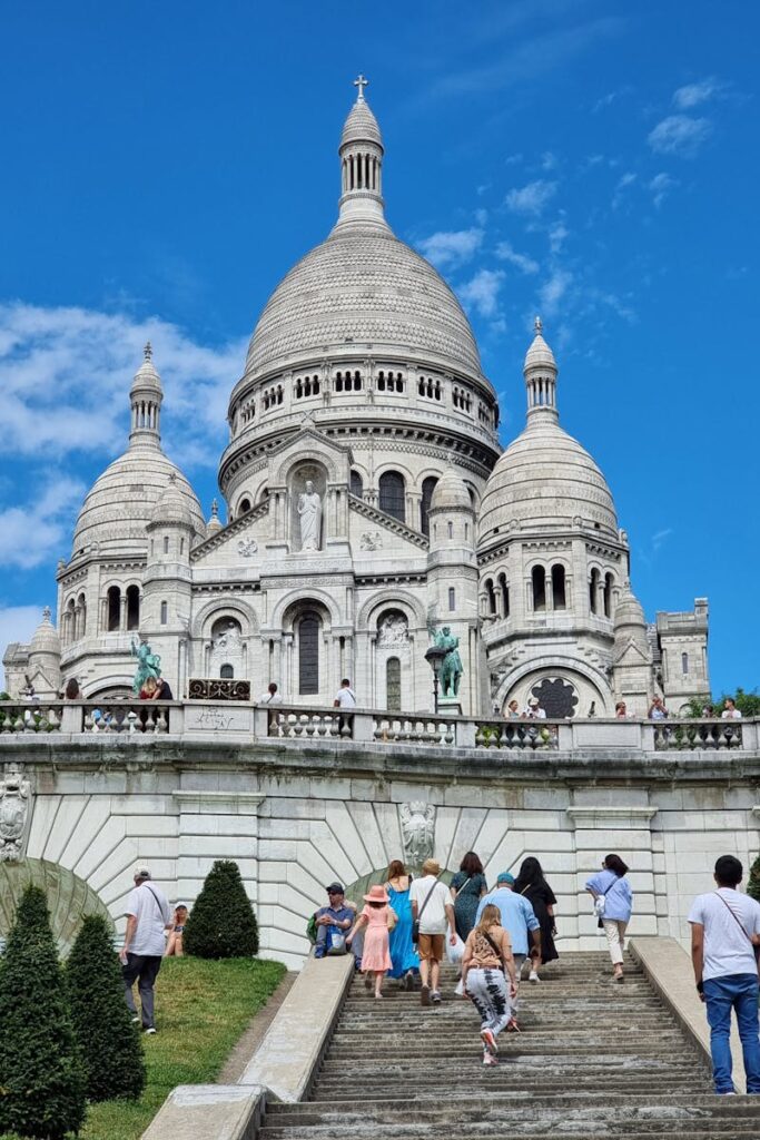 Tourists climb steps to Sacré-Cœur Basilica in Paris under a clear blue sky.